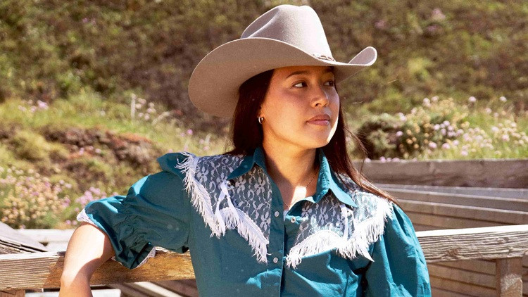 Person in a light-colored cowboy hat and blue western-style shirt leaning on a wooden railing outdoors, with trees and wildflowers in the background.