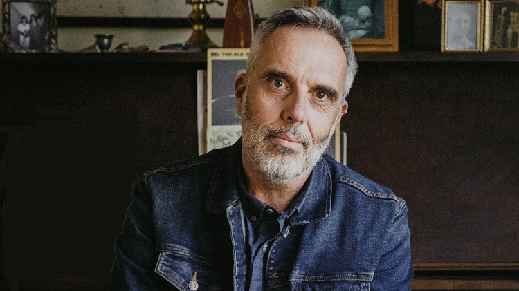 Portrait of a person with short gray hair and a beard, wearing a denim jacket and seated indoors in front of shelves displaying framed photos and books.