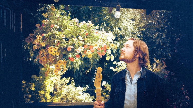Performer stands with his guitar looking up. In the background is a colourful bush in a vignette.