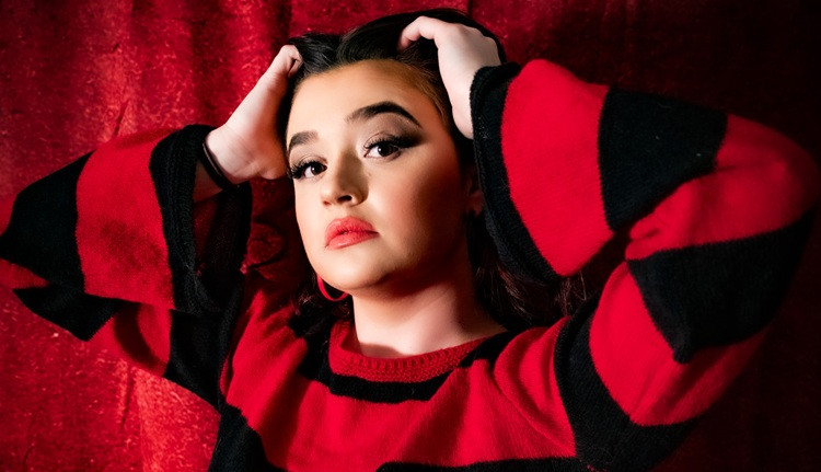 Performer poses in a red and black striped sweater, holding her head with her hands in front of a red background.
