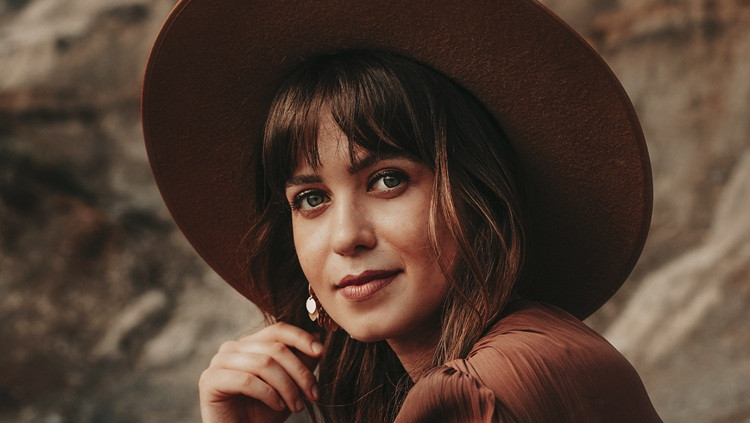 Close-up portrait of a person wearing a wide-brimmed brown hat and brown top, seated outdoors against a rocky background.