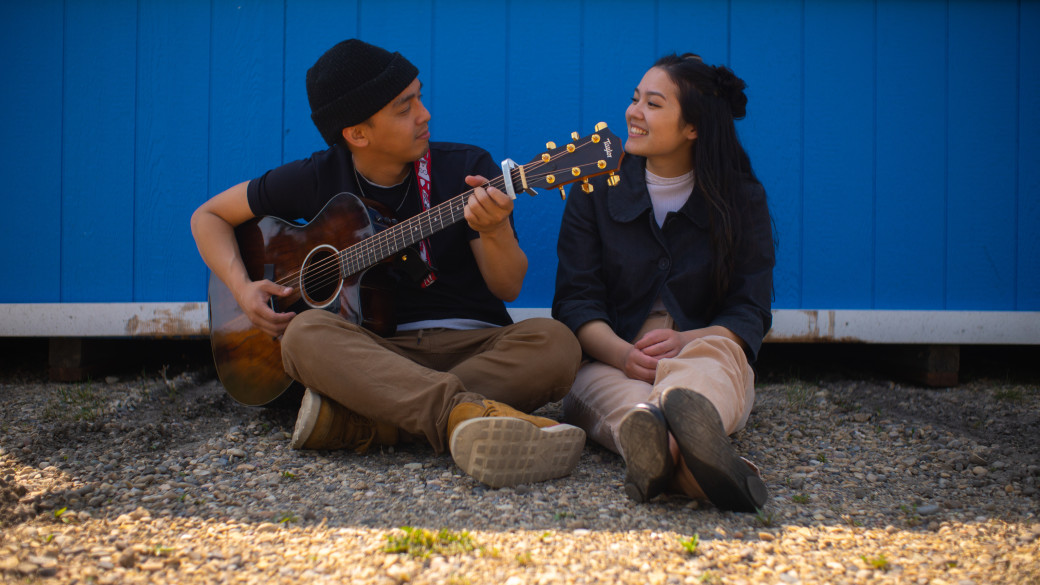 Musical duo sit together in front of blue wall singing and playing guitar.