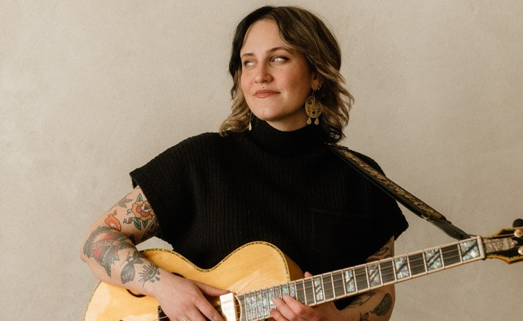 Performer stands in front of grey background holding her acoustic guitar.