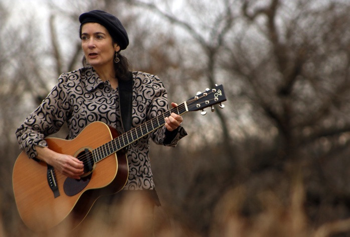 Performer holds her acoustic guitar in a field with trees in the background with no leaves.