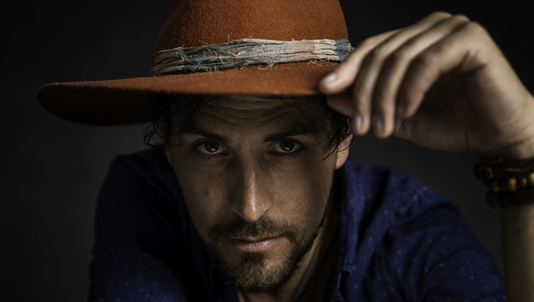 Close-up portrait of a person wearing a brown hat and dark blue shirt, looking toward the camera while adjusting the brim, against a dark background.