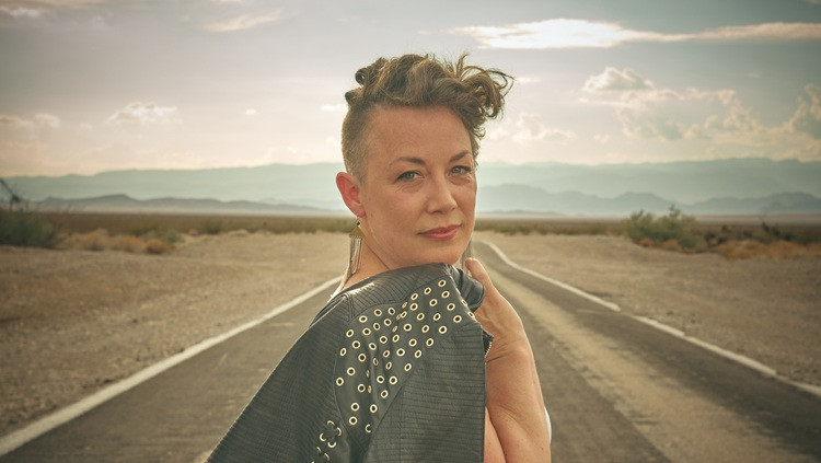Person standing on a desert road with mountains in the distance, wearing a dark jacket with metal eyelet details and looking toward the camera.