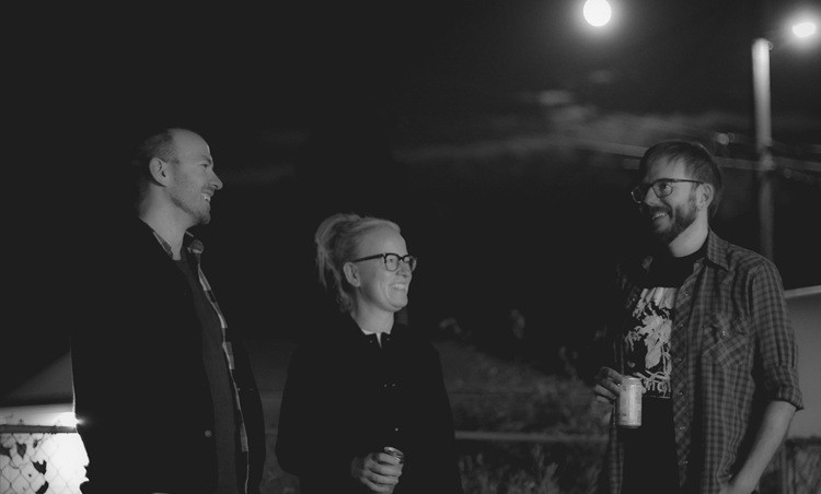 3 members of a band stand together outside under the moonlight laughing and drinking.