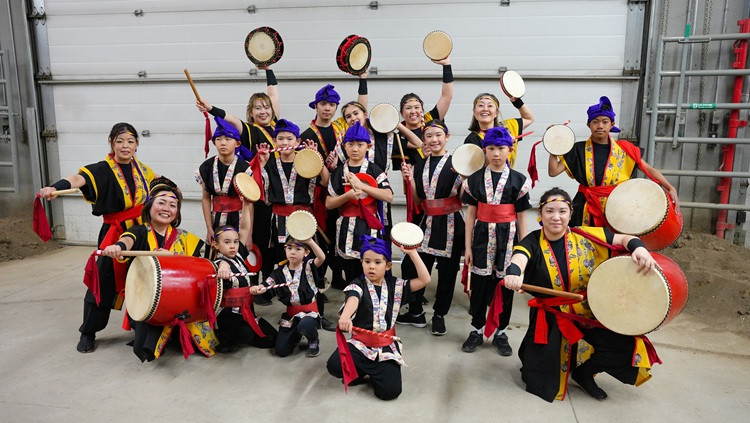 Group of performers in coordinated traditional drumming attire posing indoors with handheld drums and large barrel drums, arranged in rows and holding drumsticks and instruments for a group photo.