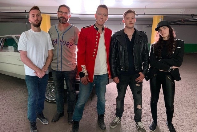 Five band members standing together in a parking garage, wearing a mix of casual and performance attire, posing in front of a vintage vehicle.