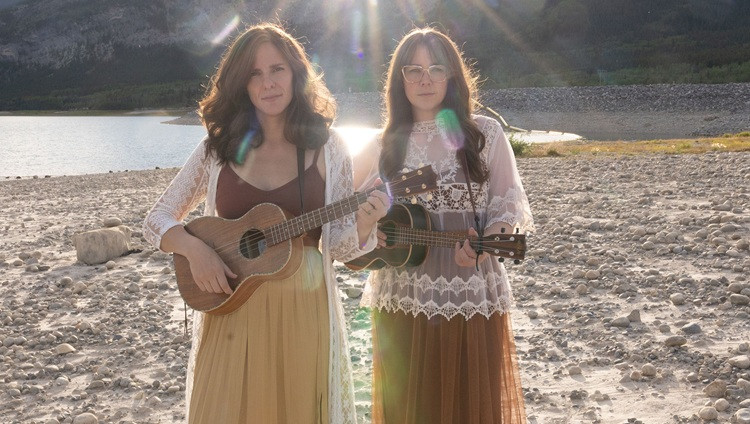 Musical duo stand on a beach with beams of sunlight shining behind them. They both hold ukuleles.