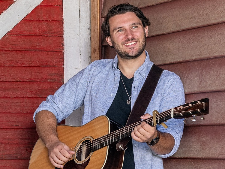 Performer leans against a wall holding his acoustic guitar.