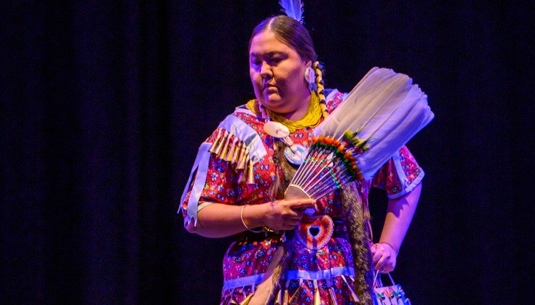 Performer in a colorful regalia-style outfit holding a decorative fan while standing on a stage under purple lighting.