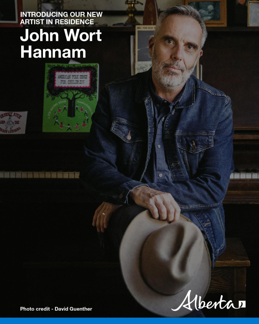 John Wort Hannam sits in front of a piano, wearing a denim jacket with a cowboy hat in his hand