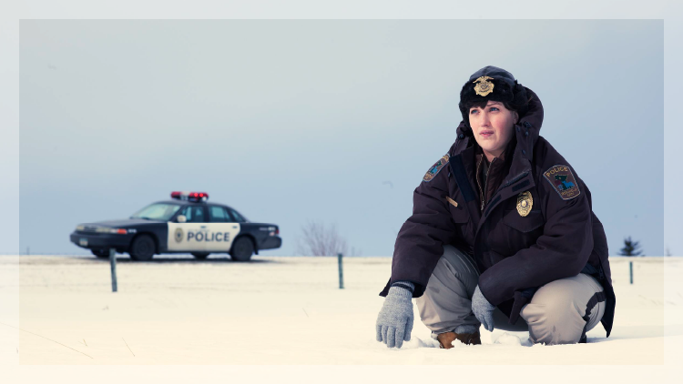 Female police officer in foreground dressed in heavy winter coat kneeling in snow with a police car in background.