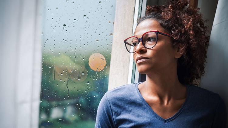A women looks out an apartment window. It's overcast and raining outside.
