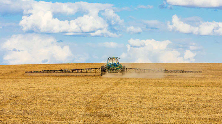 Green tractor spreading fertilizer on a yellow field under a blue, cloudy sky