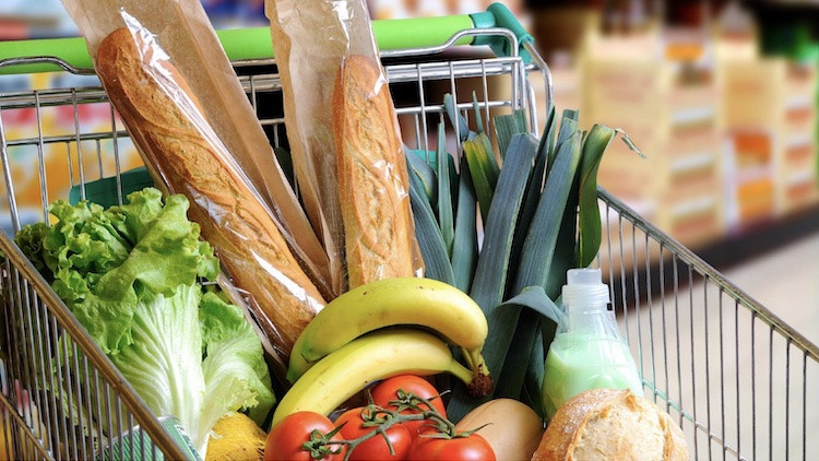 Silver metal grocery cart with a green handle filled with produce, 2 French baguettes wrapped in plastic an paper, lettuce, green onions, bananas, tomatoes