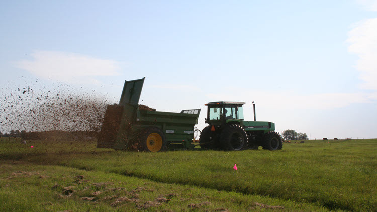 Green tractor pulling a manure spreader with manure spraying out of the back onto a green field