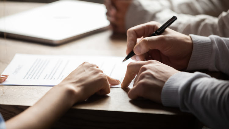 Arms and hands of 3 people sitting at a table pointing at a document for the person holding a black pen to sign