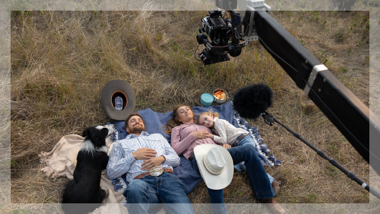 Overhead shot of man, woman, child and dog laying on a blanket on a field with a boom arm of a camera overhead.