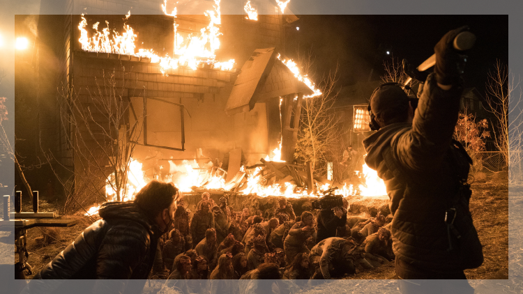 Burning house in background with a pit in foreground filled with people and a man holding a boom mic on a movie set.