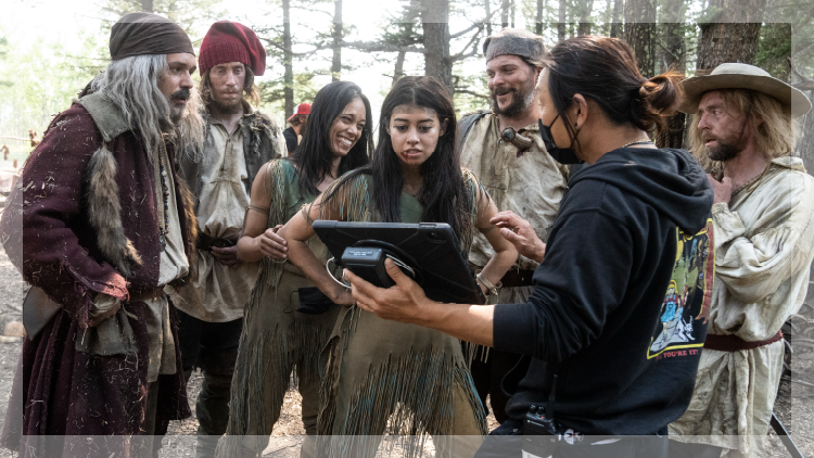 Group of people dressed in ragged clothes in a forest gathered around a tablet on a movie set.