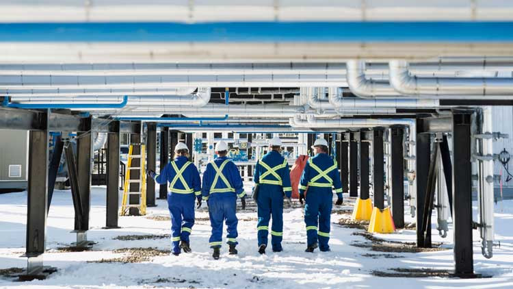 Four oil workers walk underneath a platform of pipes. They're wearing blue coveralls and white hard hats. There is snow on the ground.