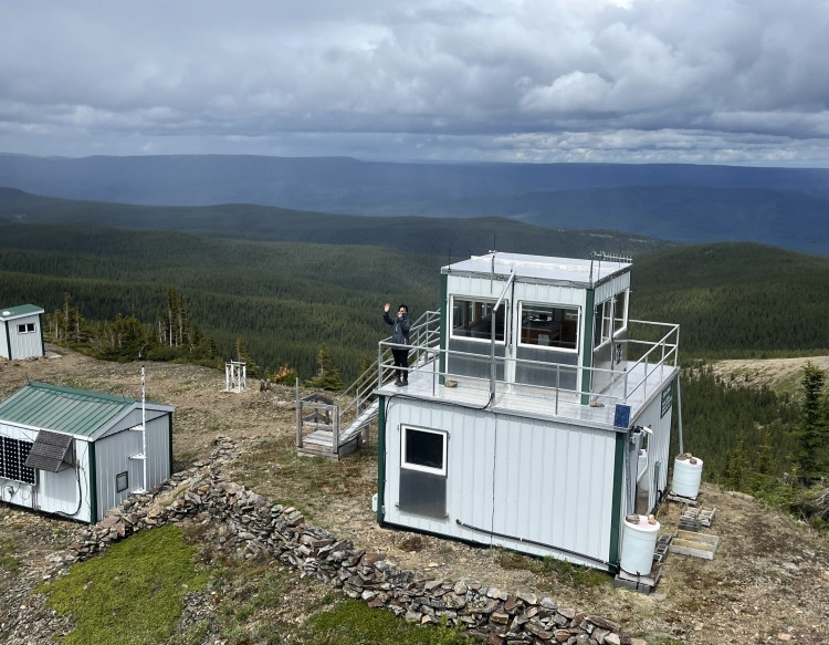 A wildfire lookout tower sits on a mountaintop surrounded by dense forest under a cloudy sky. A lookout observer stands on the stairs outside the tower, waving toward the camera. Smaller utility buildings and solar panels are nearby.