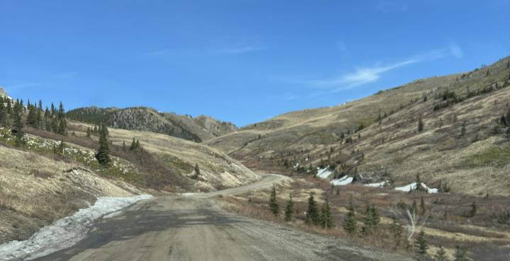 A landscape photo show snow on the edge of the road running through hills with lots of dry vegetation. 
