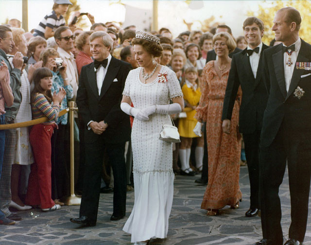 An archival photo of Queen Elizabeth the Second walking, with a crowd of people watching.