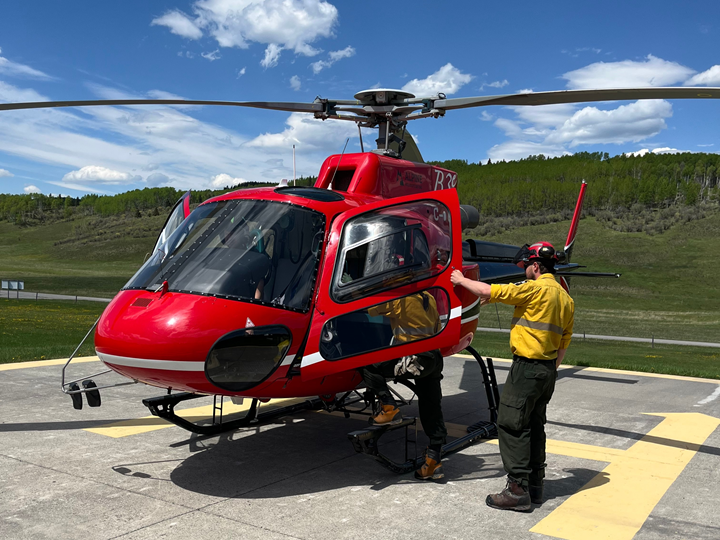 With a blue sky background, two wildland firefighters are entering a red helicopter on a cement helipad.