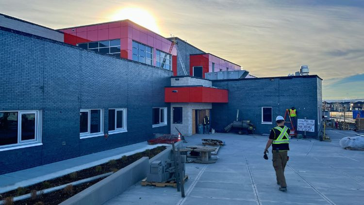 Exterior of a school under construction, showing a modern building with red and gray panels, a concrete walkway, and a construction worker walking on site in daylight.