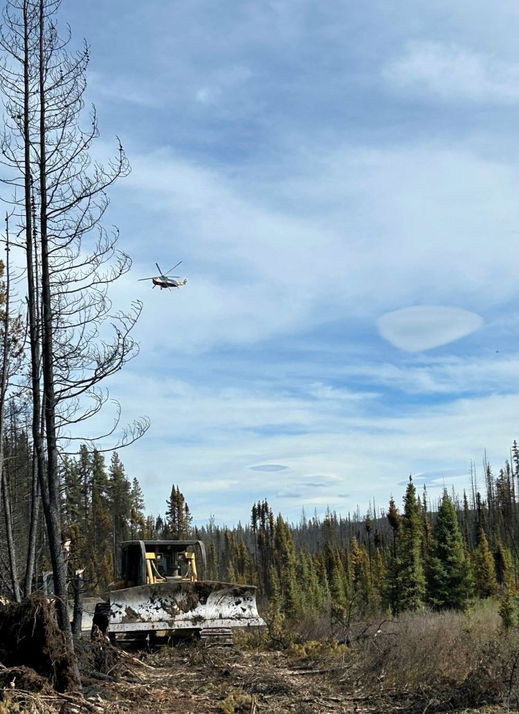 A heavy helicopter hovers over the wildfire and a dozer is parked on the containment line.