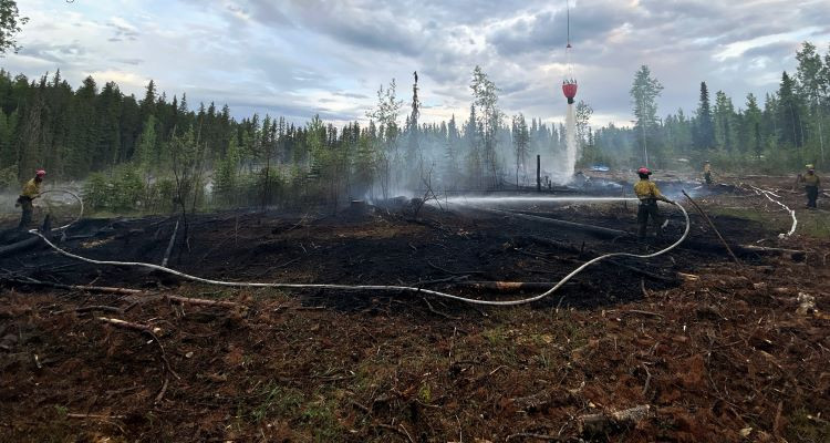 A helicopter bucket hangs over a wildfire, hose wrapping around the black with firefighters putting water on the burn.