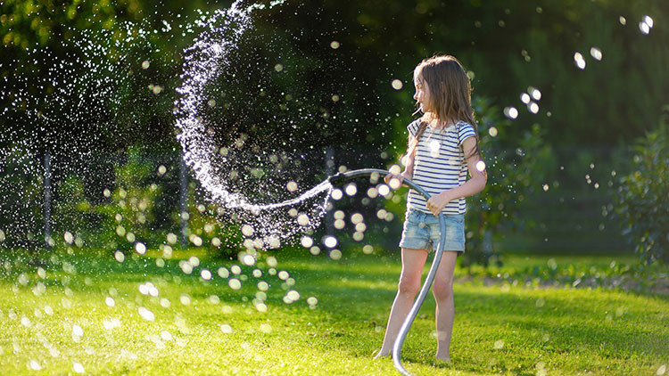 Young girl dressed in a striped t-shirt and jean shorts, standing holding a garden hose watering a green lawn