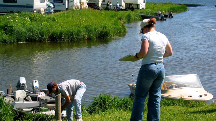 Photo of a woman with a clipboard standing at a grassy lakeside, conducting a survey with an angler.