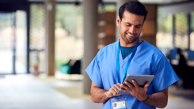 A male healthcare professional wearing scrubs looks at a tablet with a slight smile on his face.