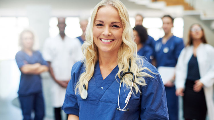 A young nurse wearing blue scrubs smiles at the camera. A group of healthcare workers stands in the background.