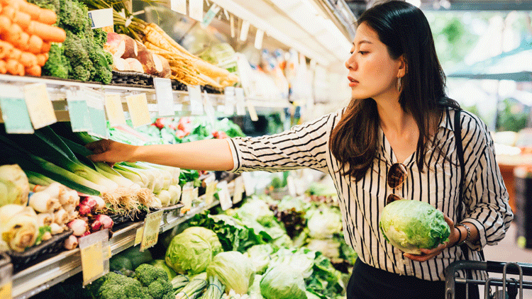 A woman shops the produce aisle in a grocery store. She has a shopping basket over her arm and holds a head of lettuce while reaching for celery.