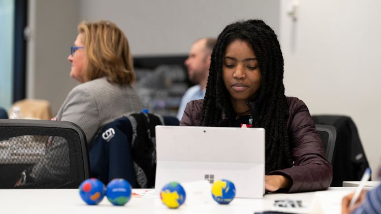 A person with long hair uses a laptop at a table in an indoor setting with others in the background.