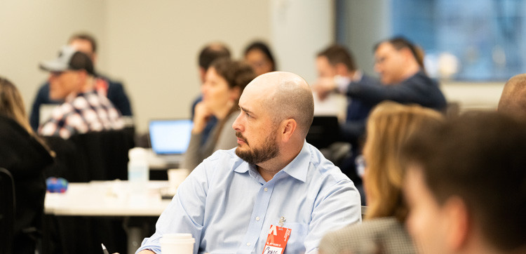 A man in a blue shirt and a beard sits at an indoor event, surrounded by seated attendees.