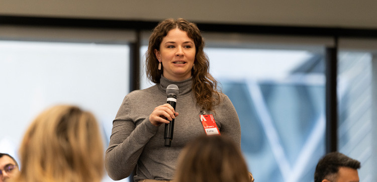 A woman standing at a presentation, holding a microphone, at a well-lit indoor event.