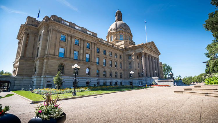 Photo of the Alberta Legislature taken from the north-east corner. It's sunny day, with blue skies. Two black planters sit in the foreground with pink flowering plants.