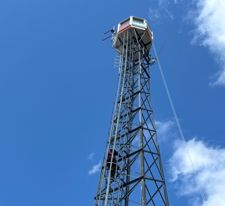 A lookout tower stretches into a bright blue sky, with a person climbing up the metal structure toward the observation cabin at the top.