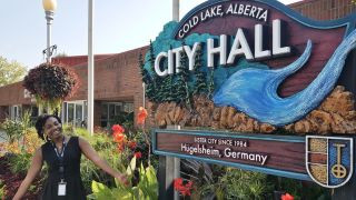 Person standing outdoors near a large decorative sign that reads “Cold Lake, Alberta City Hall” surrounded by flowers and greenery.