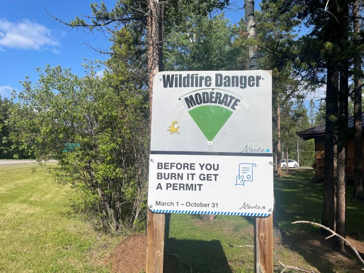 A wildfire danger sign in a forested area reads: 'Wildfire Danger: MODERATE. Before you burn it, get a permit. March 1 – October 31.' The sign includes an illustration of a document with a checkmark and the word 'Alberta' appearing twice.