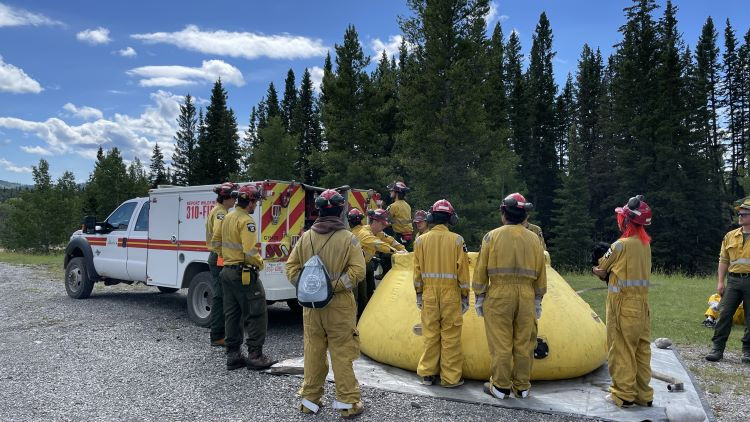In an outdoor setting surrounded by tall spruce and pine trees and a partly cloudy sky, a group of wildland firefighters in yellow uniforms are gathered with youth from the Tsuut'ina Nation. They are standing beside a large yellow water tank and a wildland firefighting truck. One firefighter is demonstrating how to refill the fire engine with water, while the youth observe and participate in the training activity.