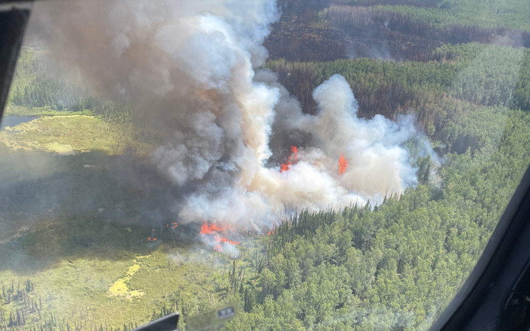 A large plume of smoke rises from a fire in a forested area, which we see aerially 