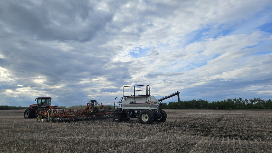 A tractor pulls an air seeder through a field of stubble. The sky is cloudy and wide overhead.