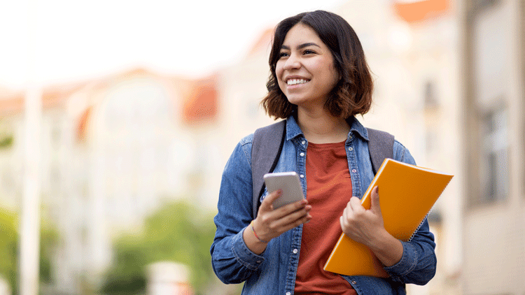 A young college student smiles and looks off to her right. She's holding her phone in one hand and some books in the other arm.